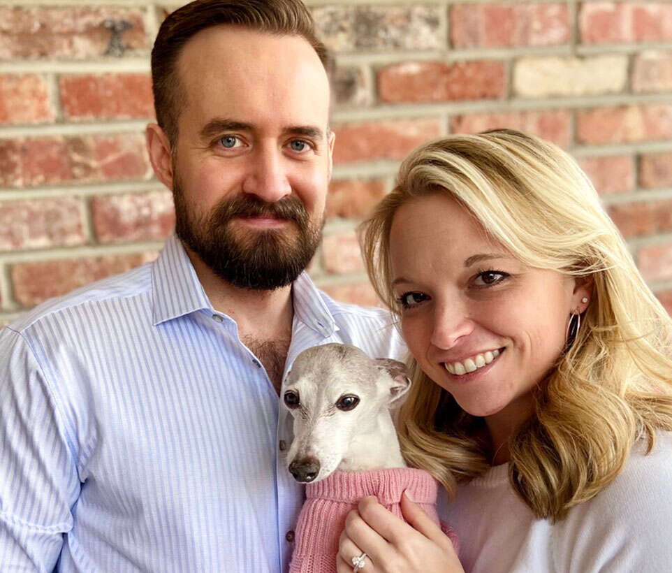 A couple stands together, smiling, while holding a small dog wearing a pink sweater.