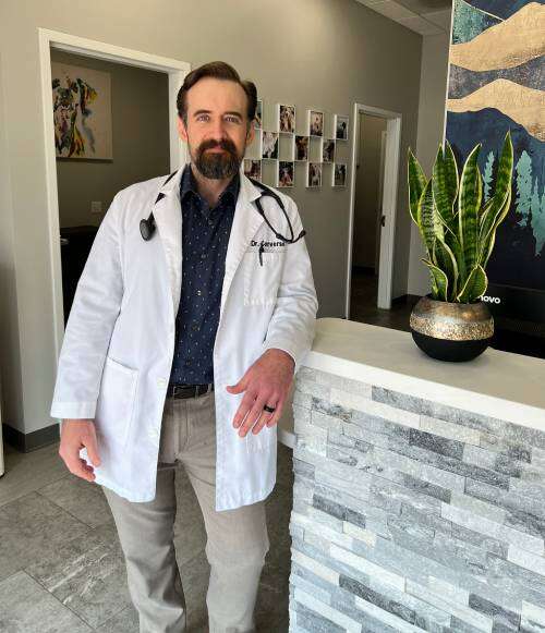 A man in a white coat with a stethoscope stands by a reception desk with a potted plant.