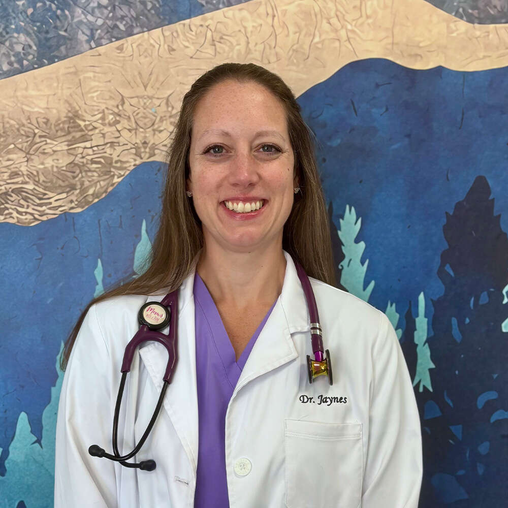 A smiling woman in a white coat with a stethoscope standing against a mountain-themed background.